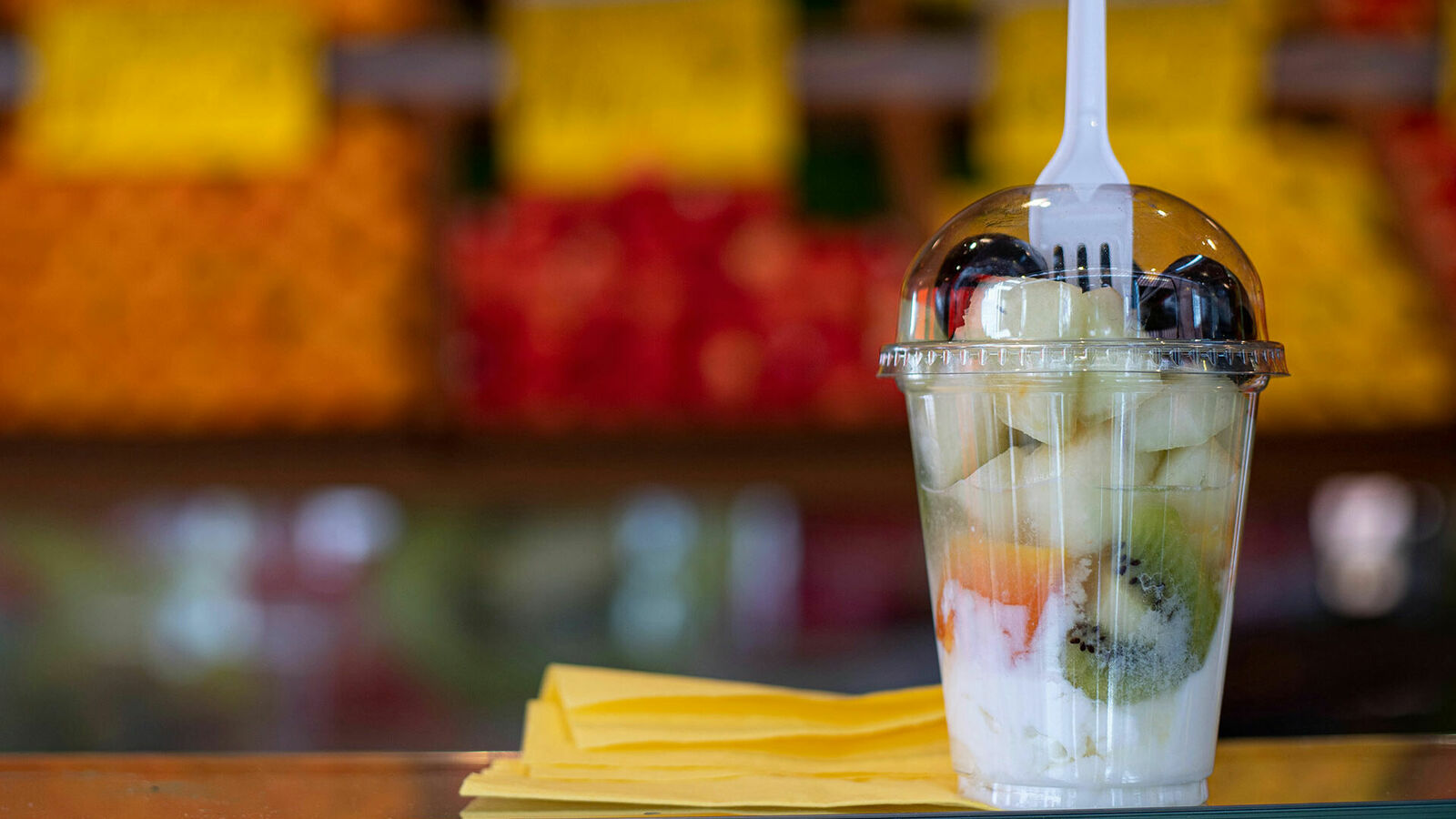 Auf einem Tisch steht ein durchsichtiger Kunststoffbecher mit Obst und Joghurt