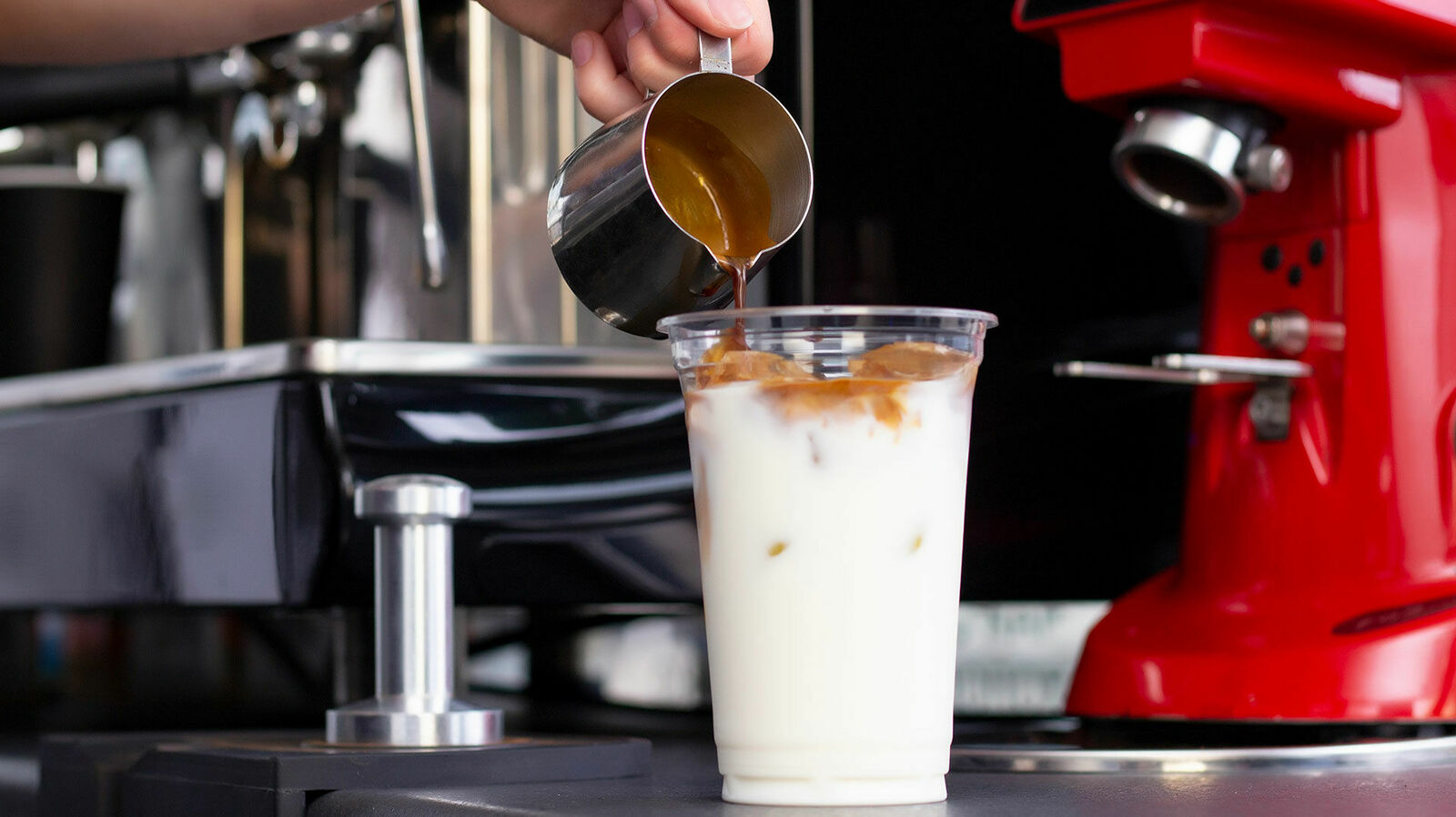 In einen Einwegbecher aus durchsichtigem Plastik, der mit Milch und Eiswürfeln gefüllt ist, wird ein Espresso hinzugegeben
