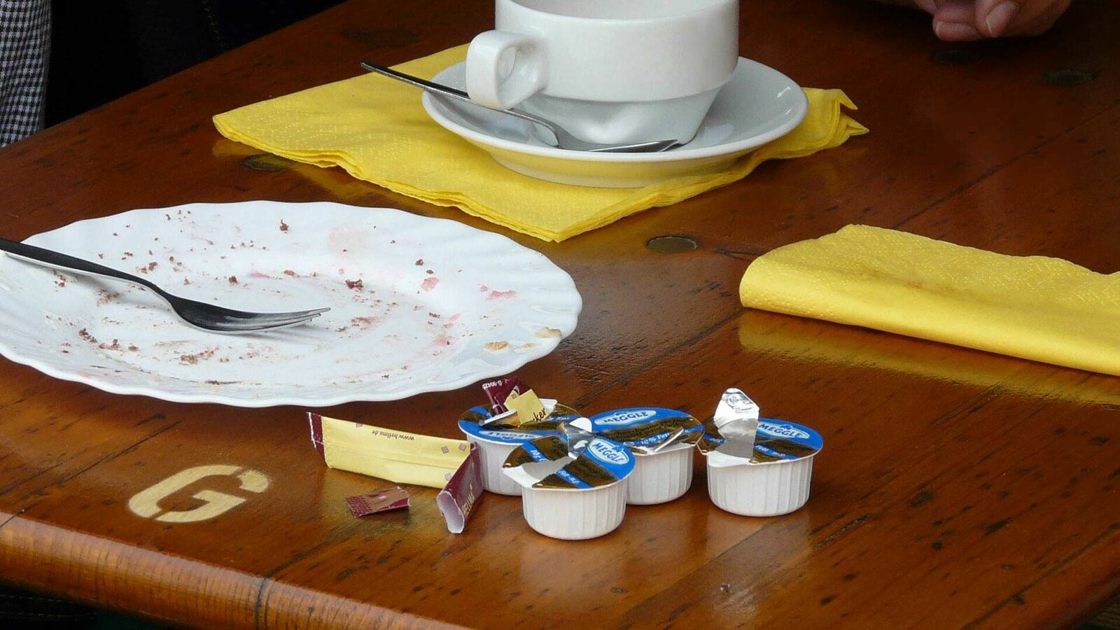 Auf einem Tisch stehen neben einer Kaffeetasse, einem leergegessenen Kuchenteller und zwei gelben Servietten vier geöffnete Mini-Verpackungen mit Kaffeesahne und ein Stick mit Zucker