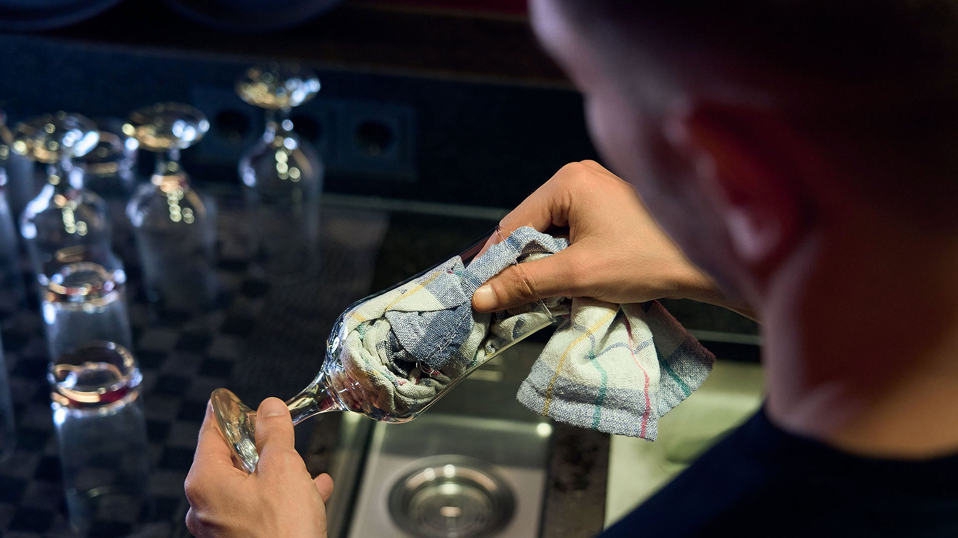 Inconvénient du lavage des verres à la main : transmission de germes Un homme se tient devant l'évier d'un comptoir de bar et sèche un verre qui vient d'être lavé avec un chiffon de polissage